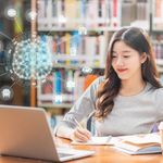 In a library, a young woman is studying with a laptop open in front of her. An AI graphic appears above the computer.