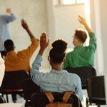 A classroom setting with students raising hands, facing the teacher. Sunlight fills the room, creating a warm, engaged learning atmosphere.