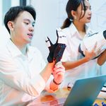 Two people in a lab, wearing white shirts, interact with robotic arms and gloves at a desk. A laptop is open, suggesting a tech-focused discussion.