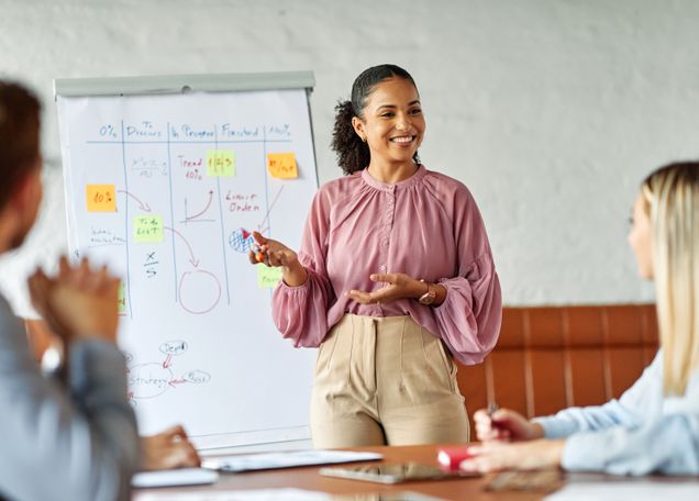 Smiling woman presents in a meeting, gesturing towards a flip chart with colorful sticky notes and diagrams. Collaborative tone.