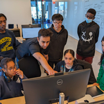 A group of students and a teacher are gathered around a computer screen in a classroom setting, focused and engaged in learning.