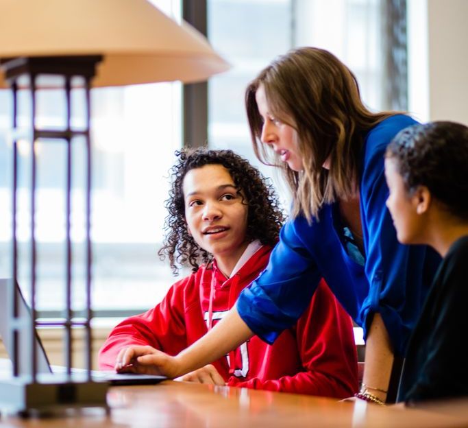 A teacher in a blue top leans over to assist two students, one in a red hoodie, at a desk with a laptop.