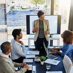 A team in a bright office. A person presents confidently in front of a screen showing graphs, while three colleagues engage thoughtfully.