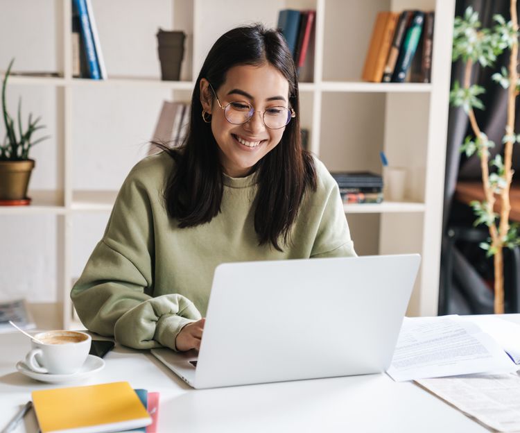 Young woman smiling and working on a laptop computer at her desk with a cup of coffee next to her.