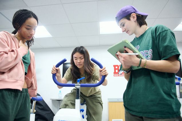 Three students in Sargent College focus on using a stationary bike in a course context. One rides, while the others observe, one with a notepad, conveying teamwork and concentration.
