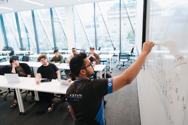 A man writes equations on a whiteboard in a bright classroom. Several students sit at desks, some with laptops, attentively watching, conveying focus.