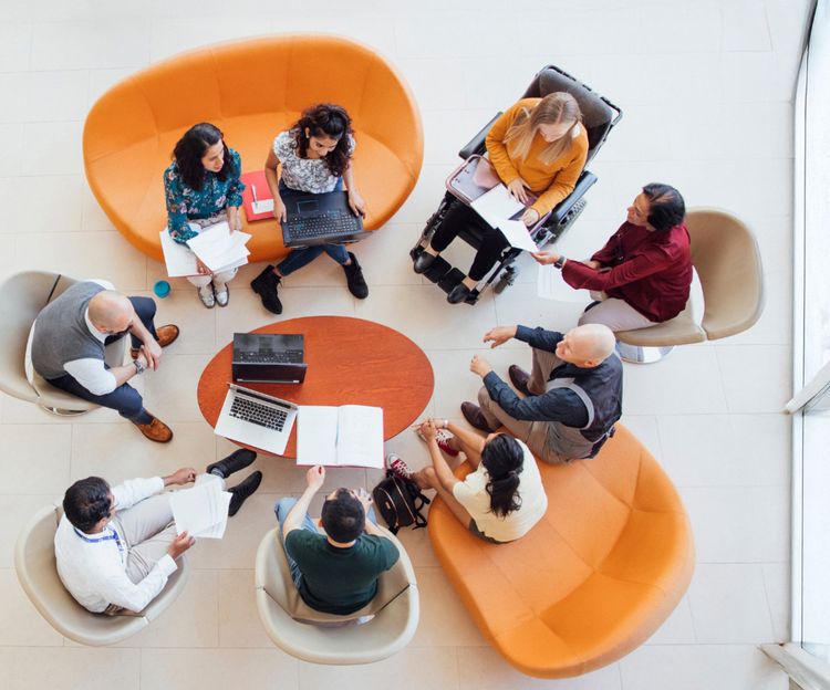 An overhead shot of a group of people sit in a circle, discussing around a table with laptops and papers. The mood is collaborative.