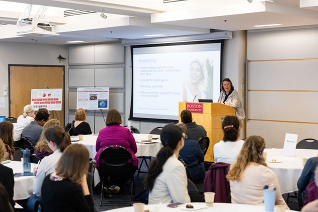 A speaker at a podium presents to an attentive audience in a seminar room. A screen displays "Humanizing" with supportive text. Emphasizes academic engagement and collaborative learning.