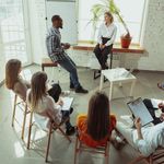 A group seated in a circle for a meeting in a bright room. One person is standing, speaking, while others take notes, fostering a collaborative atmosphere.