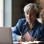 Woman with gray hair wearing headphones, smiling while writing in a notebook. She sits at a wooden table with a laptop in a cozy kitchen setting.