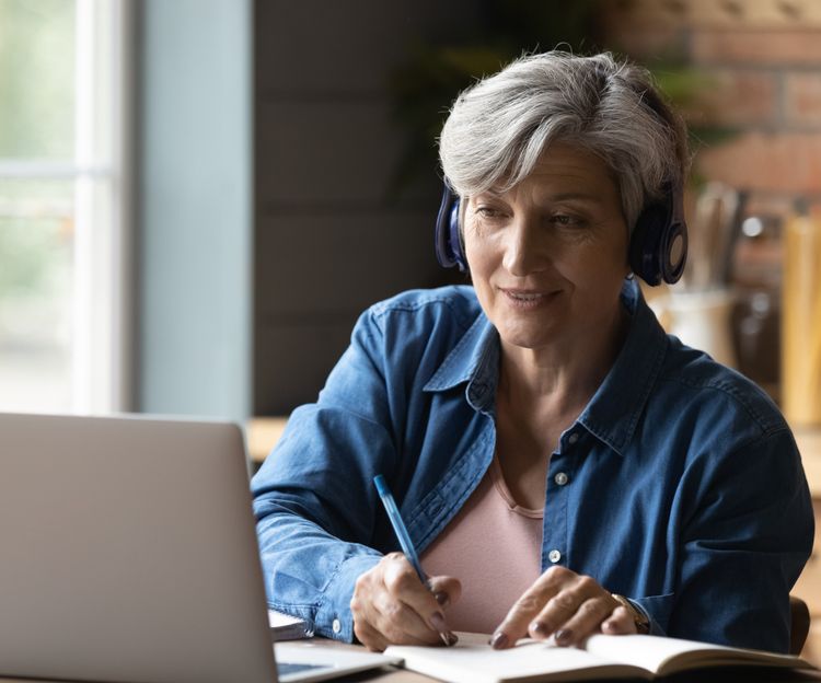 Woman with gray hair wearing headphones, smiling while writing in a notebook. She sits at a wooden table with a laptop in a cozy kitchen setting.