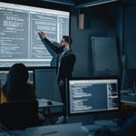 Dark classroom with a person pointing at a large screen displaying code. Students at desks focus on screens, creating a focused, tech-driven atmosphere.