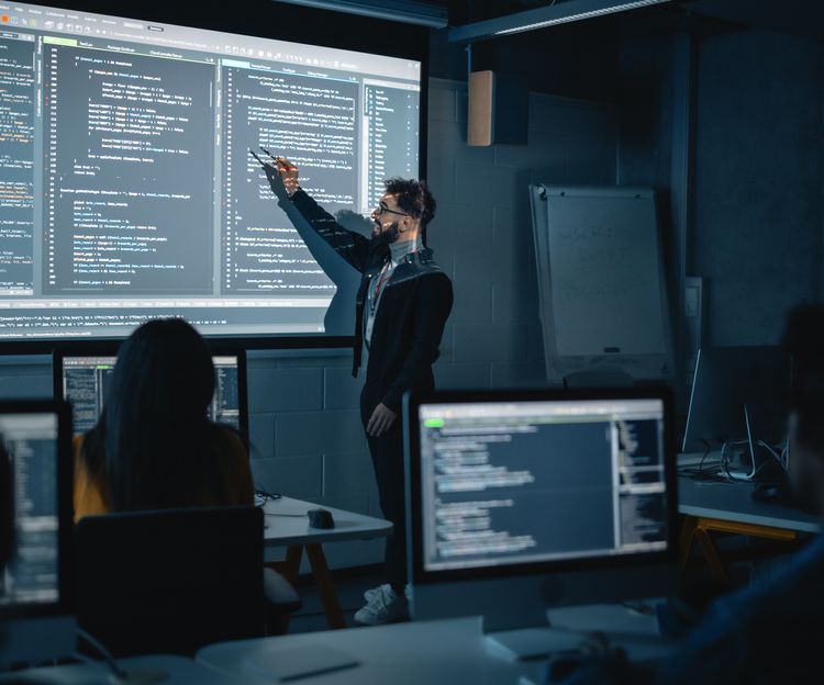 Dark classroom with a person pointing at a large screen displaying code. Students at desks focus on screens, creating a focused, tech-driven atmosphere.