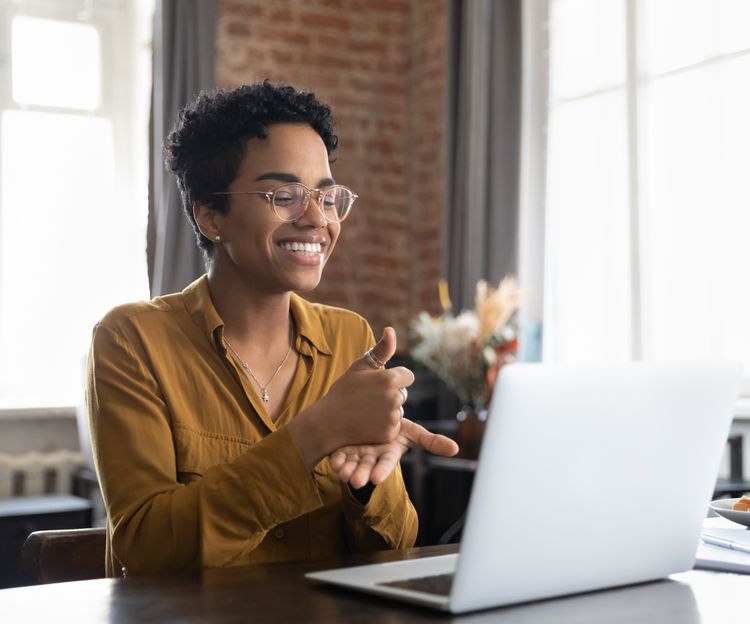 Smiling person wearing glasses and a mustard shirt using sign language during a video call on a laptop in a bright, cozy room.