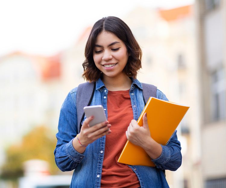 Smiling young woman holds a smartphone and yellow folder, wearing a denim jacket and backpack. Background shows blurred city buildings and trees.