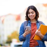 Smiling young woman holds a smartphone and yellow folder, wearing a denim jacket and backpack. Background shows blurred city buildings and trees.
