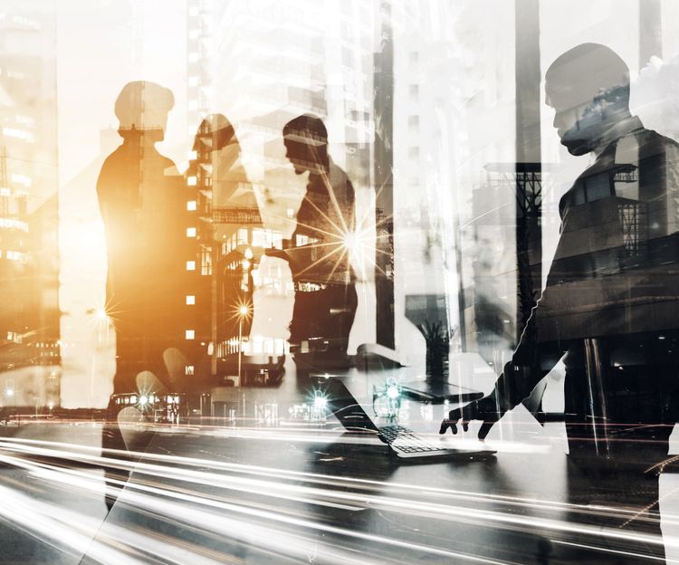 A blended image showing silhouettes of business people working on laptops overlaid with a cityscape and blurred lights, conveying a sense of dynamic teamwork and urban life.