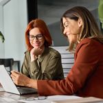 Two women in an office setting discuss work at a laptop. One has red hair and glasses, while the other wears a red blazer. Both appear engaged and smiling.