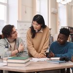 Three students collaborate in a library, discussing notes at a table with books. The atmosphere is friendly and focused, with a whiteboard in the background.