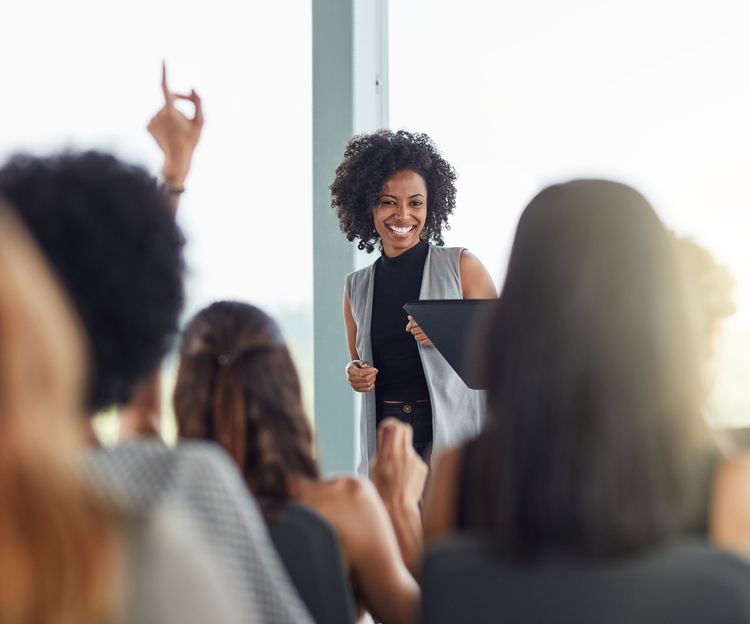 A smiling woman stands in front of an audience, holding a tablet. One attendee raises a hand, suggesting engagement. The atmosphere is bright and lively.