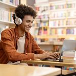 Dedicated smiling male student engaged in online learning at college library, using laptop to study, surrounded by bookshelves