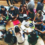 A group of students sits around a circular table, engaging in lively discussion. They have notebooks, tablets, and papers, conveying collaboration.