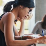 Close up shot of young woman writing notes with classmates studying in background. Students learning in college library.
