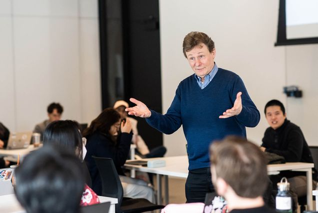 Man in blue sweater gestures while speaking to a classroom of attentive students. The room is bright and modern, conveying an engaging atmosphere.