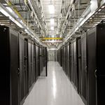 Long corridor of a data center with rows of black server racks on either side, overhead cables, and bright lighting, conveying efficiency and modernity.
