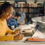 A woman in a yellow sweater smiles while studying at a library. She writes in a notebook beside a laptop, with bookshelves and others working in the background.