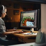 A person wearing headphones works intently on digital art at a computer. The screen displays a colorful landscape design. The setting is cozy and focused.