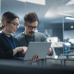 A man and woman in a modern office collaborate over a laptop. They appear focused and engaged, with a blurred background of tech equipment visible.