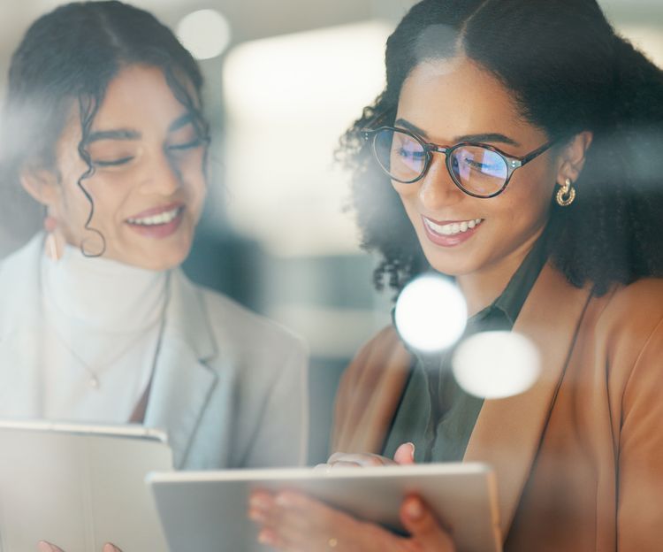 Two women smiling and collaborating in an office. One in a light blazer, the other in glasses and a brown jacket. Both are looking at a tablet. Bright atmosphere.