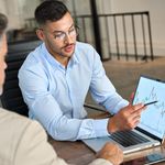 Two men in an office setting discuss a financial chart on a laptop. One points at the screen, conveying analysis and collaboration.