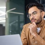 A man with glasses and a contemplative expression works on a laptop. He wears a brown shirt, with modern office lighting in the background.