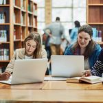 A group of four young adults studies together in a bright library, focused on laptops and books, conveying a collaborative and studious atmosphere.