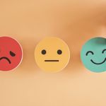 Three emoji faces on a peach background: red sad face, yellow neutral face, and a blue happy face being picked up, conveying varied emotions.