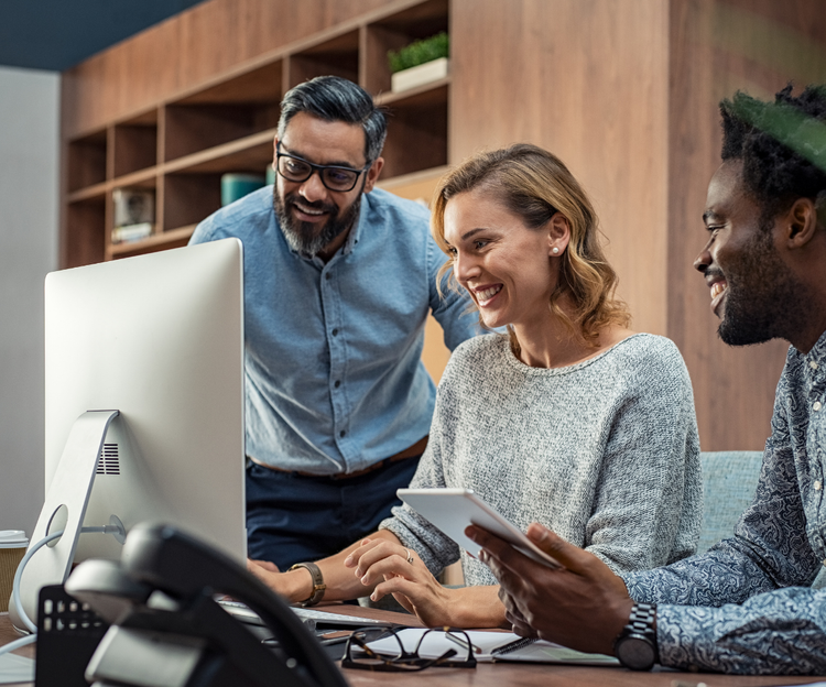 A group of three people work together and look at something on a computer monitor. The atmosphere is joyful and collaborative.