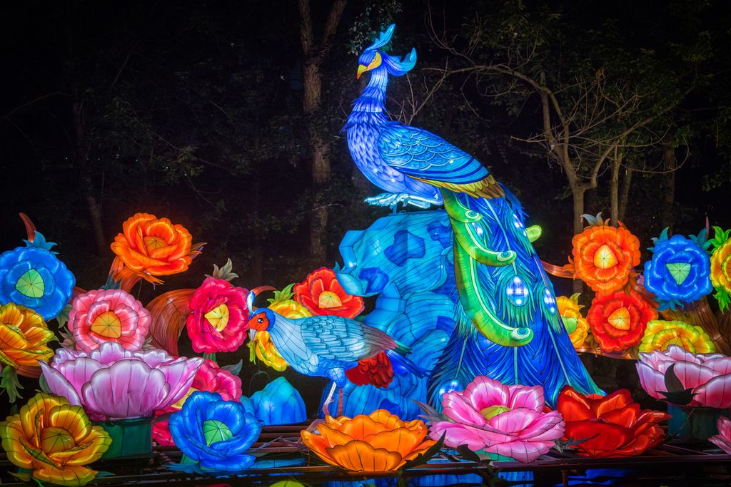 A lantern shaped like a peacock is illuminated in blue light with other colorful flowers and plants lit up beside the bird