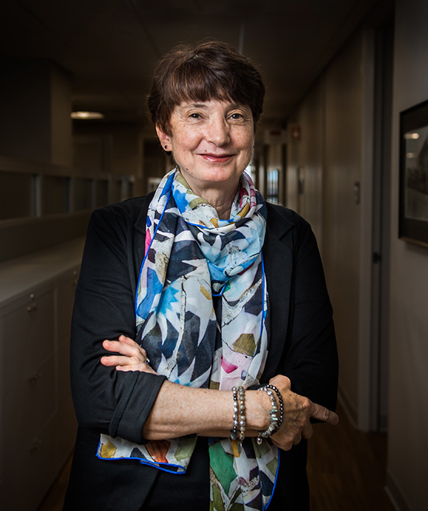 Photo: Julie Palmer, a white woman with short dark brown hair and wearing a long sleeved black shirt and light blue, light grey, and black patterned silk scarf, smiles and poses with armed crossed in a dark, dimly lit hallway. She is illuminated and shown clearly.