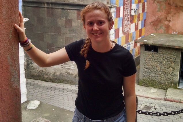 Photo: Student researcher Elizabeth Hannigan, a white woman with long red hair tied into a braid and wearing a black tee shirt and jeans, poses at an archaeological site.