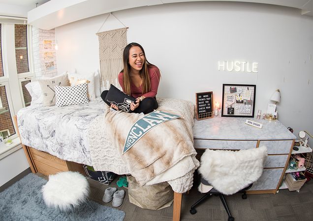 A female Boston University student sits crosslegged on her Myles Standish Hall dorm room bed laughing.