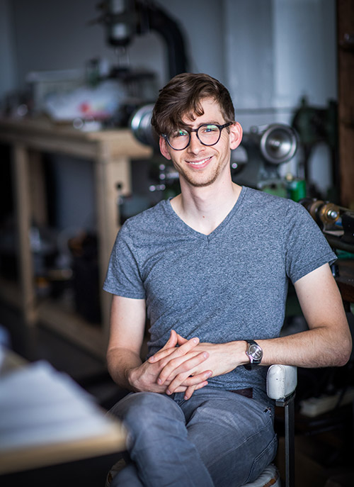 Schon Horology founder Ian Schon poses for a sitting portrait in his Allston mechanical engineering and design studio. Tools used for manufacturing can be seen in the background.