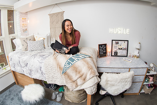 A female Boston University student sits crosslegged on her dorm room bed laughing.