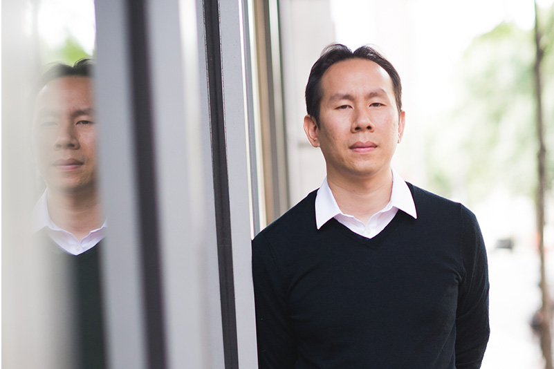 Portrait of Jerry Chen, Boston University College of Arts and Sciences assistant professor of biology, leaning against windows outside a building so that his reflection can be seen to the left.