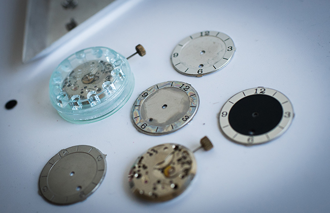 Disassembled watch faces and watch movements lay on a white table in Ian Schon's Schon Horology studio in Allston, MA.