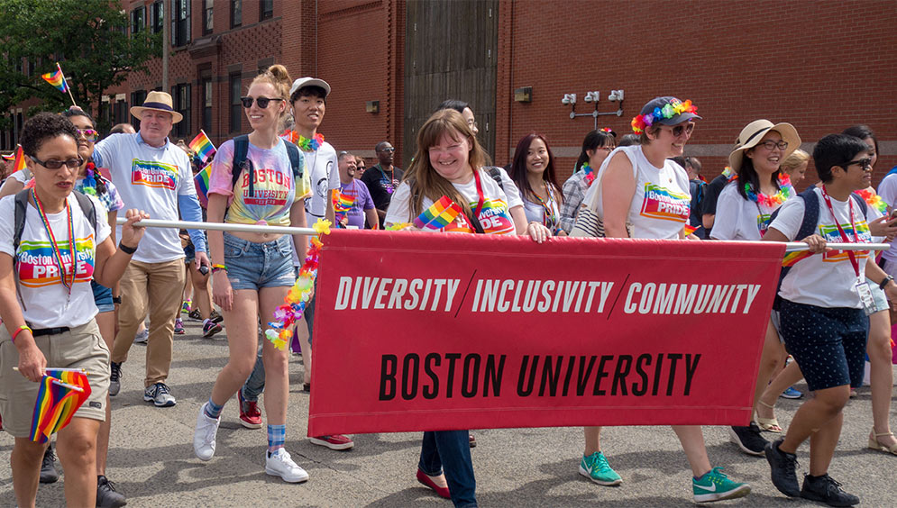 Boston University faculty and staff members march in the Boston Pride parade wearing t-shirts with the slogan 'Boston University Pride' and holding a banner with the slogan 'Diversity Inclusivity Community, Boston University'.