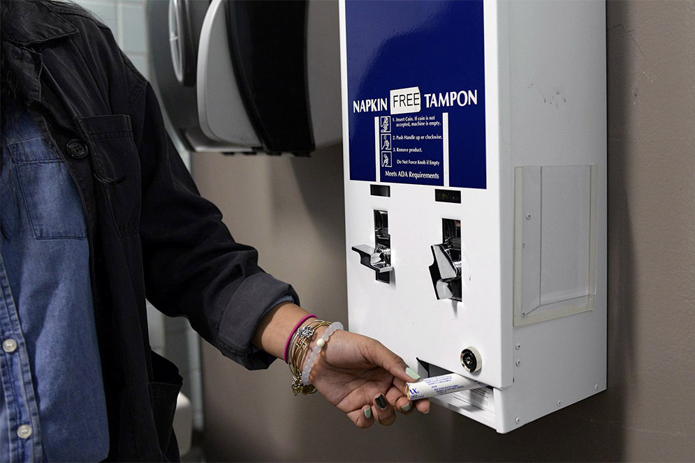 A young woman removes a tampon from a blue and white free tampons dispenser in a restroom.