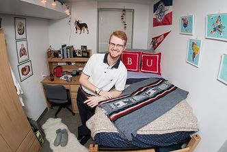 BU student Evan Dann stands smiling in his dorm room.