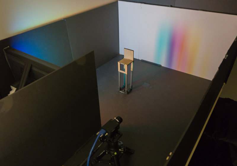 A rainbow penumbra on a far wall is created by a bright scene displayed on an LCD monitor (left) and a chair (center).